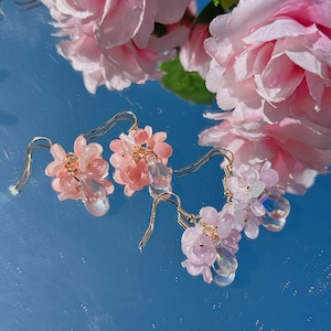 May include: A pair of dangle earrings with pink and purple flower-shaped charms and clear teardrop crystals. The earrings are made with gold-toned metal.