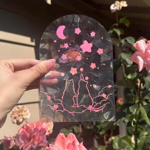 May include: A clear glass panel with a mosaic design features two pink silhouettes of foxes looking up at a crescent moon and pink stars. The panel is held in front of a pink rose bush.