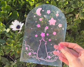 May include: A clear iridescent sticker with a black cat silhouette reaching up to pink stars and a crescent moon. The sticker is on a green leafy background.
