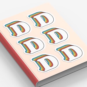 May include: A collection of white stickers featuring the letter "D" in a retro style. Each letter has a layered effect with blue, yellow, and red outlines. The stickers are arranged on a light pink notebook.