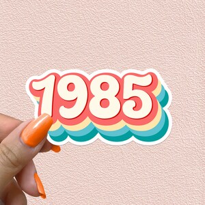 May include: A sticker with the year 1985 in a retro rainbow font. The numbers are white with a red, yellow, blue, and green outline.