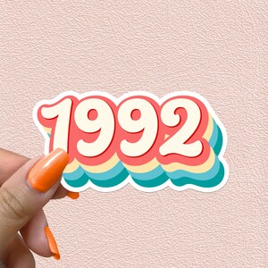 May include: A sticker with the year 1992 in a retro style font. The numbers are white with a red outline and are layered on top of a rainbow of colors, including blue, yellow, and pink.