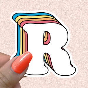 May include: A die-cut sticker featuring the uppercase letter R in a bold, white font with a thick black outline. The left side and inner curves of the letter are accented with a layered, retro-inspired rainbow stripe pattern in blue, yellow, orange, and pink.