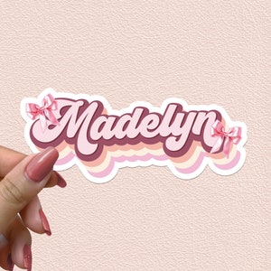 May include: A pink and white sticker with the name "Madelyn" in a retro font. The name is surrounded by pink and white stripes and two pink bows.