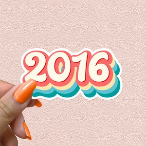 May include: A sticker featuring the year 2016 in a retro rainbow font. The numbers are white with a red outline and are layered on top of a blue, yellow and pink background.
