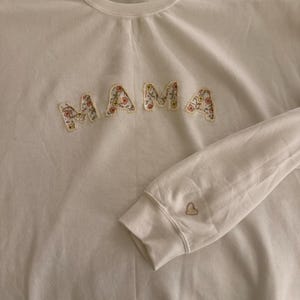 May include: White sweatshirt with the word "MAMA" in floral fabric letters across the chest. A small embroidered heart is on the left sleeve. The sweatshirt is hanging on a white hanger.
