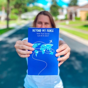 May include: A person holding a blue journal with the title "Beyond My Fence: World Travel Journal (with 50 US states)" and a world map with a plane on it.