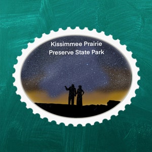 Puede incluir: Una pegatina ovalada blanca con un borde festoneado, que presenta la silueta de dos personas de pie bajo un cielo estrellado. El texto "Kissimmee Prairie Preserve State Park" está impreso en negro en la parte superior de la pegatina.