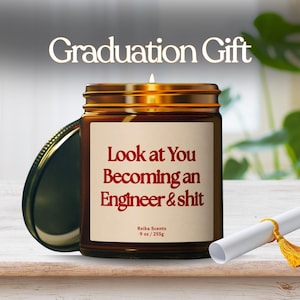 Puede incluir: Una vela encendida en un frasco de vidrio marrón con tapa dorada. La etiqueta dice "Look at You Becoming an Engineer & shit". El texto "Graduation Gift" está encima del frasco. Un diploma enrollado con una borla dorada está en primer plano.