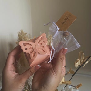 Puede incluir: Una caja de regalo rosa en forma de mariposa con una pequeña botella de vidrio con un tapón de corcho dentro de una bolsa de organza blanca. La bolsa tiene una etiqueta marrón con el nombre "Kayleigh" escrito en ella.