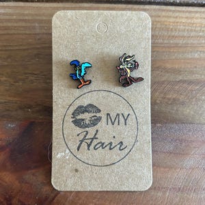 Puede incluir: Un par de pendientes con personajes de dibujos animados. Los pendientes están hechos de metal y presentan un correcaminos azul y un coyote marrón. Los pendientes están en una tarjeta marrón con el texto "MY Hair" impreso en ella.