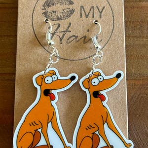 May include: A pair of dangle earrings featuring a cartoon dog with a surprised expression. The dog is orange and white and has a long tongue sticking out. The earrings are silver and have a hook closure. The earrings are on a brown card with the text "MY Hair" printed on it.