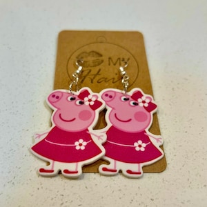 May include: A pair of Peppa Pig earrings in pink and white. Each earring depicts the cartoon pig wearing a pink dress and a bow. The earrings hang from silver-coloured hooks and are presented on a brown card with the text "My Hair".