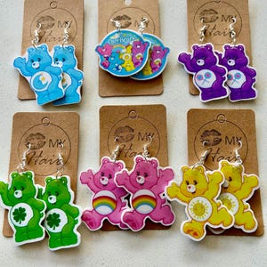 May include: Care Bears earrings in various colours, including blue, purple, green, pink, and yellow. Each pair features a different Care Bear design with silver-tone ear wires. The earrings are displayed on brown cardboard earring cards.