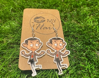 Op de afbeelding: Een paar zilveren oorbellen met cartoonfiguren van Mr. Bean. De oorbellen zijn op een bruine kaart met de tekst "MY Hair" erop gedrukt.