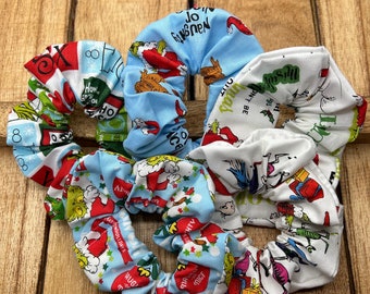 Puede incluir: Tres scrunchies coloridos con temática navideña. Los scrunchies están hechos de tela con una variedad de patrones, incluyendo un scrunchie azul con un patrón de Grinch, un scrunchie blanco con un patrón de Grinch y un scrunchie rojo y verde con un patrón navideño.