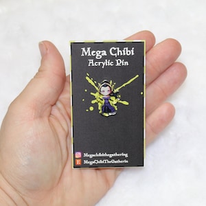 May include: A black card with white text that reads "Mega Chibi Acrylic Pin". A yellow paint splatter design is behind a small, colourful, anime-style character pin.