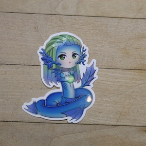 May include: A blue and green cartoon sticker of a mermaid with long hair holding a green orb. The mermaid has a blue tail and fins.