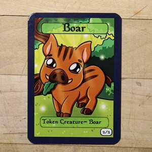 May include: A cartoon illustration of a brown boar with a green leaf in its mouth. The card is green and blue with the text "Boar" and "Token Creature - Boar 2/2".