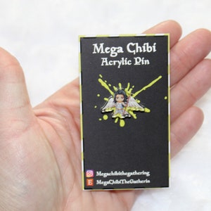 May include: A black card with white text that reads "Mega Chibi Acrylic Pin". A colorful acrylic pin of a chibi angel with yellow wings is attached to the card. The background of the card is black with yellow paint splatters.