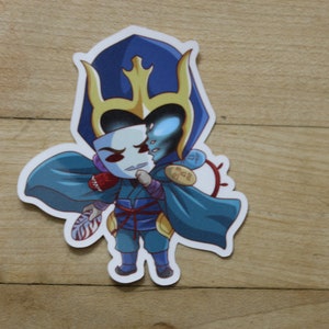 Puede incluir: Un personaje azul y dorado con una máscara, sosteniendo un bastón de caramelo rojo y blanco. El personaje lleva una capa azul y una corona dorada.