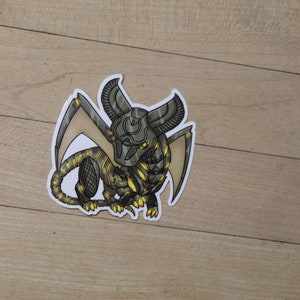 May include: A black and gold metallic dragon sticker with wings outstretched. The dragon has a detailed design with intricate patterns and a helmet.