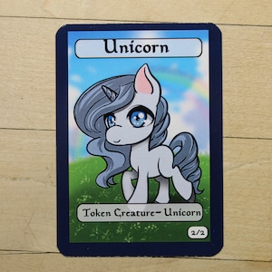 May include: A white unicorn with a blue horn and blue eyes. The unicorn is standing on a green field with a rainbow in the background. The card reads "Unicorn" and "Token Creature - Unicorn" with a power and toughness of 2/2.