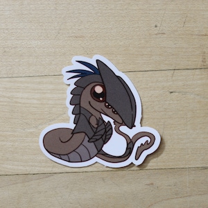 May include: A cartoon sticker of a small, gray, and blue creature with large eyes. The creature is curled up with its tail in its mouth.
