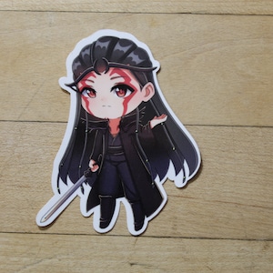 Puede incluir: Una pegatina en blanco y negro de un personaje de anime chibi con cabello negro largo, vistiendo un atuendo negro y sosteniendo una espada. El personaje tiene marcas rojas en la cara.