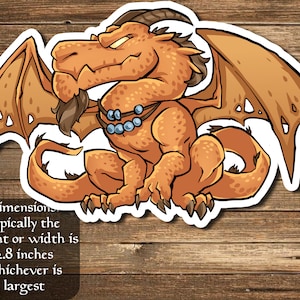 May include: A cartoon orange dragon with blue beads around its neck and a brown beard. The dragon has wings and a tail. The image is on a wood background. Dimensions typically the height or width is 2.8 inches whichever is largest.