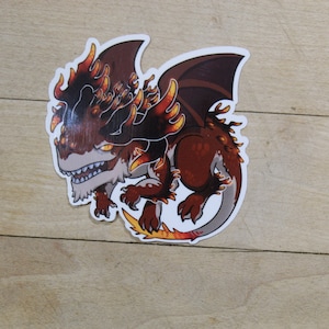 May include: A sticker featuring a cartoon dragon with a fiery mane and wings. The dragon is primarily red and brown, with yellow and orange accents. It has a white belly and a toothy grin. The sticker is on a light brown wooden surface.
