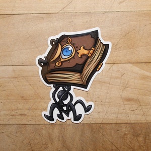 Könnte beinhalten: Ein braunes Buch mit einem einzigen blauen Auge auf dem Einband, das auf vier schwarzen Metallbeinen steht. Das Buch hat goldene Akzente und einen abgenutzten, alten Look.