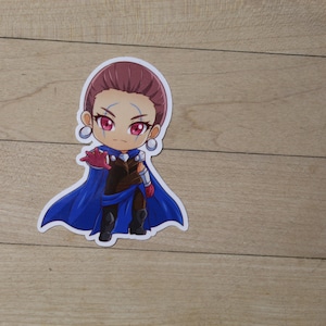 May include: A sticker of a cartoon character with purple hair, blue eyes, and a blue cape. The character is wearing a brown outfit and red gloves. The sticker is on a wooden surface.