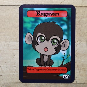 May include: A cartoon illustration of a monkey on a trading card. The card is blue with a red border and the text "Ragavan" in black. The monkey is brown with big green eyes and a surprised expression. The card text reads "Token Legendary Creature - Monkey 2/1".
