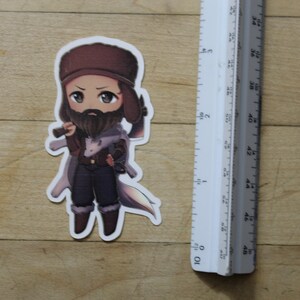 May include: A cartoon sticker of a person wearing a brown hat and a brown beard, holding a small axe. The person is wearing a brown and white jacket and brown pants. The sticker is on a wooden surface.