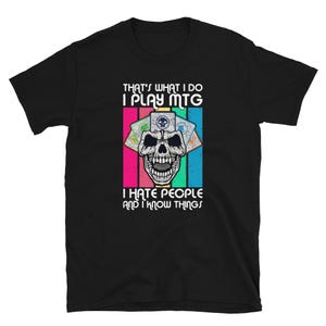 May include: Black t-shirt with a graphic featuring a skull, playing cards, and the text "THAT'S WHAT I DO I PLAY MTG I HATE PEOPLE AND I KNOW THINGS." The design includes pink, blue, green, and white stripes.