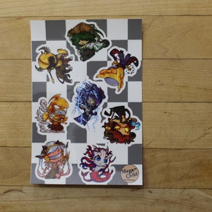 May include: A sheet of stickers featuring various cartoon characters in a chibi style. The stickers are on a black and white chequered background. The characters include a yellow and red creature, a blue and purple creature, a green creature, a red creature, a yellow creature, and a pink creature. The text "Mega-Chibi" is at the bottom of the sheet.