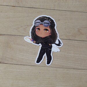 May include: A sticker featuring a cartoon illustration of a person with brown skin, black hair, and black clothing. The person is wearing goggles and holding a flaming object. The background is a light wood surface.
