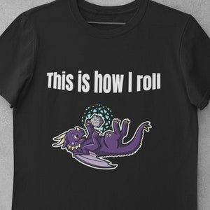 May include: Black t-shirt with white text that reads "This is how I roll". A purple dragon with wings is holding a grey and white dice.