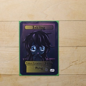 Puede incluir: Una carta morada y negra con un personaje de dibujos animados con ojos grandes y cabello largo y negro. La carta dice "Inkling" y "Criatura ficha - Inkling" con el texto "Vuelo" y "2/1".