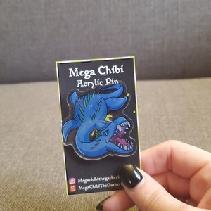 May include: A blue and green acrylic pin featuring a cartoon dragon with its mouth open. The pin says "Mega Chibi Acrylic Pin" and includes the social media handles "Megachibithegatherer" and "MegaChibiTheGatherer".