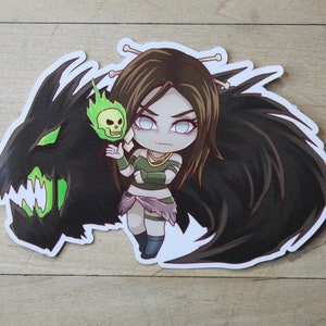 Op de afbeelding: Een sticker met een cartoonvrouw met lang bruin haar, die een groene en bruine outfit draagt en een groen-zwarte schedel met een groene vlam vasthoudt. Ze staat voor een zwart en groen wezen met vleugels.