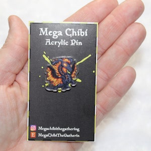 May include: A black card with the text "Mega Chibi Acrylic Pin" and a small, colorful, dragon-like creature on a yellow and green splatter background.
