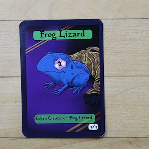 May include: A purple card with a green border and gold accents. The card features a cartoon illustration of a blue frog-like creature with a single eye. The text on the card reads "Frog Lizard" and "Token Creature - Frog Lizard 3/3".
