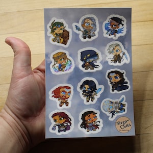 Puede incluir: Una hoja de pegatinas con ilustraciones de personajes de The Gathering en estilo chibi. Las pegatinas están sobre un fondo azul con nubes blancas. Los personajes llevan todos atuendos diferentes y tienen expresiones diferentes. El texto "Mega-Chibi The Gathering" está en la parte inferior de la hoja.