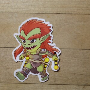 May include: A green and red cartoon goblin sticker with a fierce expression, wearing armor and holding two yellow and black staffs.