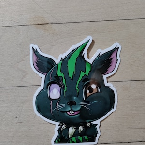 May include: A black and green cartoon sticker of a creature with a mischievous expression. The creature has a green mohawk, a white eye, and a brown eye.