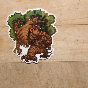 May include: A sticker featuring a cartoon tree creature with brown bark, green foliage, and orange eyes. The tree has claw-like hands and feet. The sticker is on a wooden surface.