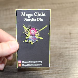 Może przedstawiać: Czarna karta z białym tekstem, który brzmi "Mega Chibi Acrylic Pin". Do karty przymocowana jest kolorowa akrylowa spinka z postacią z kreskówki. Spinka jest otoczona żółtymi plamami farby.