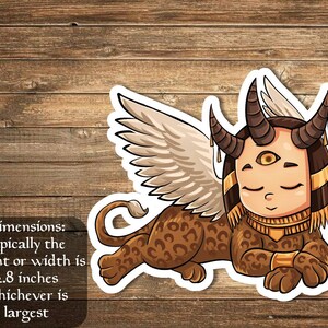 Puede incluir: Un león alado marrón y dorado con cabeza humana y un tercer ojo. El león está acostado con las alas extendidas. El león mide aproximadamente 2,8 pulgadas de alto o de ancho.